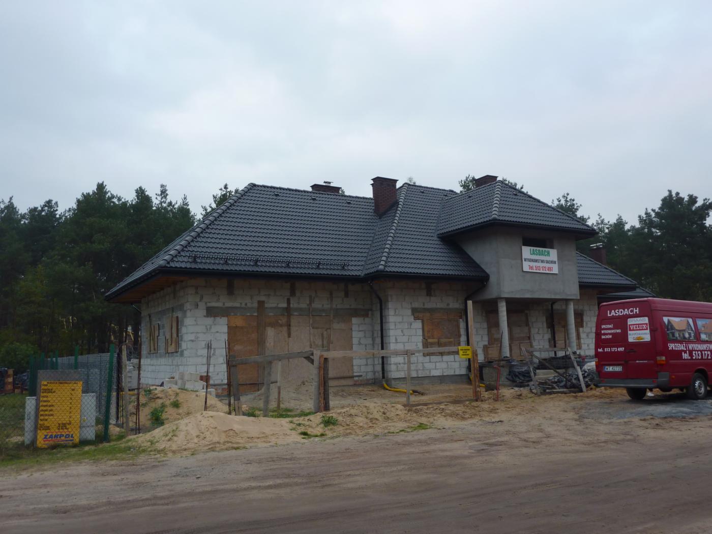 Dom w trakcie budowy z nowym, grafitowym dachem z dachówki ceramicznej, częściowo zamurowane okna, widoczny samochód dostawczy firmy dekarskiej i materiały budowlane.